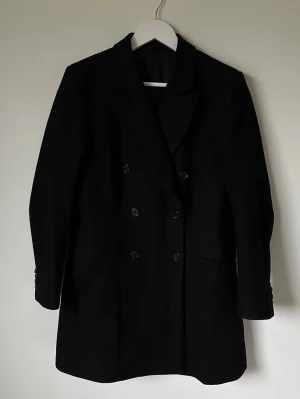 Svart kappa från J.Lindeberg - Svart kappa från J.Lindeberg med dubbelknäppning och klassisk krage. Tillverkad i 80% ull och 20% polyamid med foder i polyester. Snygg, stilren och tidlös modell som passar perfekt till höst och vinter. Fickor fram och knappar vid ärmslut. 6Q4