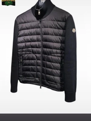 Moncler cardigan - Säljer en svart pufferjacka från Moncler med quiltad framsida och släta ärmar. Jackan har dragkedja framtill, ståkrage och diskret Moncler-logga på ärmen. Materialmix med polyester och stickade detaljer ger en clean och modern look.