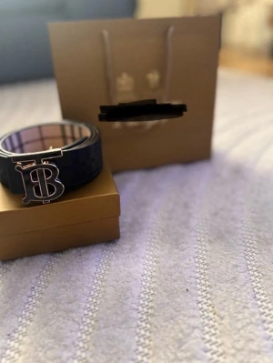 B bälte  - Snygg blå rutig bälte  från Burberry med ett stort metallspänne i form av ett B framtill. Västen har ett klassiskt rutmönster på insidan och är perfekt för dig som gillar exklusiva detaljer och stilren design. Två färger utsidan är svart o insidan är beige man kan ändra på bältet 