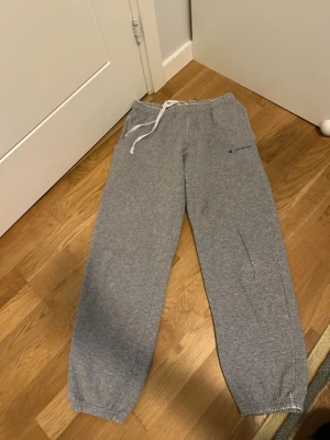 Grå mjukisbyxor från Champion - Ett par välanvända gråa Champion sweatpants med resår i midjan och bensluten. Byxorna har en dragsko i midjan och en broderad Champion-logga på vänster ben. Det finns fickor på sidorna.  Välanvända och har skavanker, se bilder. Men håller ett tag till :)