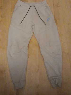 Beige mjukisbyxor från Nike med snörning - Säljer ett par beige mjukisbyxor från Nike i storlek S. Byxorna har svart snörning i midjan, ribbade muddar vid bensluten och en blå Nike-logga på vänster sida. Perfekta för chill eller träning, tillverkade i mjukt bomullsmaterial.