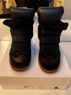 Svarta sneakers Isabel Marant med kilklack - Säljer ett par svarta sneakers från Isabel Marant med kilklack och platt gummisula. Skorna har flera breda kardborreband, detaljer i mocka och skinn samt perforerad tå. Insidan är ljusbeige och yttersulan är brun. Perfekt för dig som gillar statement-skor med cool design.