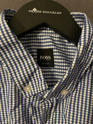 Blårutig slim fit skjorta från Hugo Boss - Snygg blårutig skjorta från Hugo Boss i slim fit-modell. Skjortan har klassisk krage, vita knappar och lång ärm. Tillverkad i ett mjukt bomullstyg som känns skönt mot huden. Perfekt för dig som gillar stilrena och tidlösa plagg. Priset kan diskuteras!