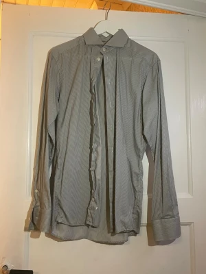 Gråvit randig skjorta från Eton - Snygg gråvit skjorta från Eton i slim fit med smala ränder och klassisk krage. Skjortan har långa ärmar med breda manschetter och knappar framtill. Perfekt för dig som gillar stilrena och moderna plagg.