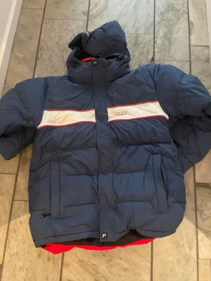 Blå Peak Performance dunjacka - Säljer en blå pufferjacka från Peak Performance med vit och röd detalj över bröstet. Jackan har huva, dragkedja och är långärmad. Perfekt för kalla dagar och har en sportig vibe med sin tjocka, quiltade design.