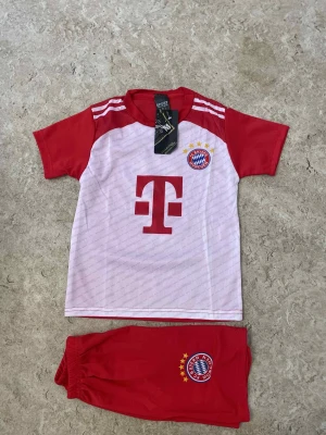 Bayern München fotbollströja och shorts - Säljer ett set med Bayern München fotbollströja och matchande röda shorts. Tröjan är vit med röda ärmar och har klubbens emblem samt sponsortryck på bröstet. Shorts i rött med klubbmärke. Perfekt för dig som älskar fotboll och vill visa ditt stöd för laget.