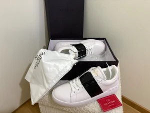 Valentino Garavani Open sneakers vit/svart - Snygga Valentino Garavani Open sneakers i vitt skinn med bred svart rem över mitten och guldfärgad logga på plösen. Platt sula med mönstrad undersida och klassisk snörning. Kommer med originalkartong och dustbag.