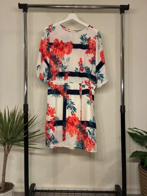 Blommig kortklänning från Holly & Whyte - Oanvänd, snygg kortklänning med blommigt mönster i rött och blått på vit botten. Klänningen har midjeband. Perfekt för sommarens alla tillfällen och ger en fräsch och elegant look. 