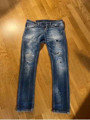 Blå dondup jeans med färgstänk - Säljer ett par blå jeans med slitningar, hål och vita färgstänk för en riktigt edgy look. Jeansen har smal passform, klassiska fem fickor och knappgylf. Materialet är jeans i bomull och de har en cool tvättad finish.