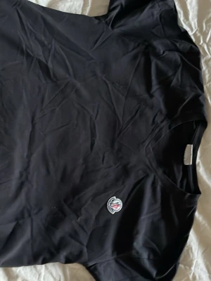 Svart t-shirt från Moncler, S - Svart t-shirt från Moncler i storlek S med klassisk v-ringning och korta ärmar. På bröstet sitter Monclers ikoniska logopatch som ger en exklusiv vibe. Materialet känns mjukt och lätt, perfekt för en chill och stilren look.