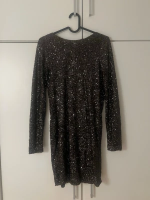 Svart glittrig paljettklänning - Säljer en svart kort klänning med glittriga paljetter över hela tyget. Klänningen har lång ärm och en riktigt snygg öppen rygg med draperad effekt. 