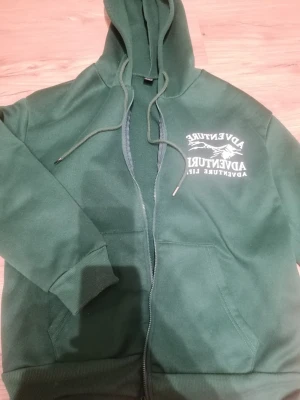 Mörkgrön hoodie med dragkedja  - Säljer en mörkgrön hoodie med dragkedja på framsidan, & har även två fickor där framme. Den har ett tryck vid bröstet. Den är lite noppig vid armarna, men inget man märker!☺️