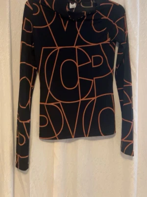 Svart långärmad polotopp med grafiskt mönster - Säljer en svart polotopp från H&M med långa ärmar och ett coolt grafiskt mönster i orange. Toppen har en tight passform och är gjord i mjuk polyester som sitter skönt mot kroppen. Perfekt för dig som gillar statement-plagg med unik design.