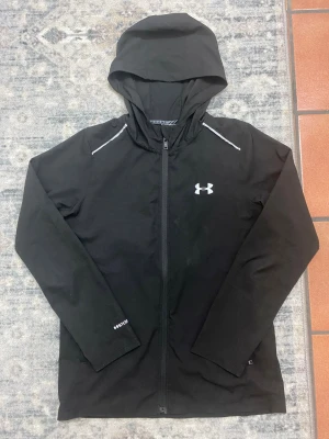 Under armour Windbreaker - Passar för både storlek S och M.  Svart vindjacka från Under Armour med huva och hel dragkedja framtill. Jackan har vita detaljer på axlarna och logga på bröstet samt stort tryck på ryggen. Tillverkad i lätt polyester som andas, perfekt för löpning och träning utomhus.