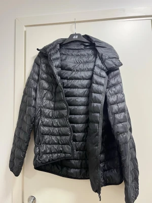 Svart pufferjacka med huva från Everest  - Svart pufferjacka från Everest med huva och horisontella quiltade partier. Jackan har dragkedja framtill och två sidofickor. Materialet är syntet och jackan är långärmad med en loose passform. Perfekt för kyliga dagar.