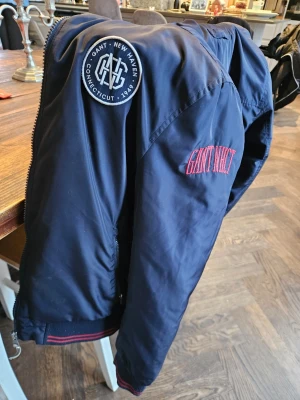 Mörkblå bomberjacka från Gant - Snygg mörkblå bomberjacka från Gant med broderad logga på ärmen och patch på axeln. Jackan har quiltad insida, dragkedja och ribbade muddar med röda detaljer. Perfekt för dig som vill ha en stilren och sportig look. 13/14 År 158/164Cm