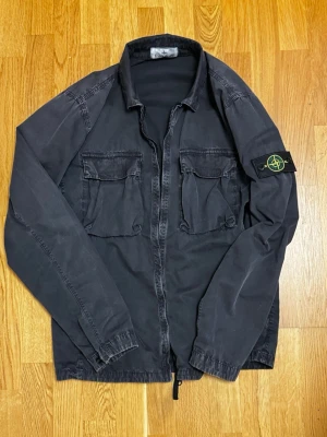 Stone Island Overshirt - Unik overshirt från Stone Island i en sliten mörkgrå färg, med två stora bröstfickor och den ikoniska patchen på ärmen. Jackan har dragkedja framtill och är tillverkad i 100 % bomull. Original låda och kvitto medföljer.
