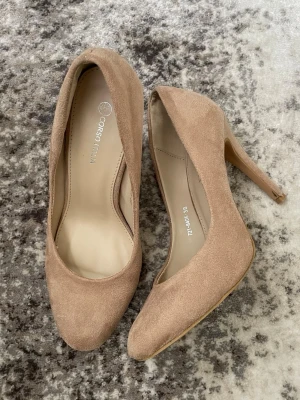 Beige pumps från Corso Italia, strl 38 - Snygga beige pumps från Corso Italia i storlek 38. Skorna har en spetsig tå och hög klack, tillverkade i mocka för en mjuk och lyxig känsla. Perfekt för dig som gillar stilrena och klassiska skor med en elegant touch.
