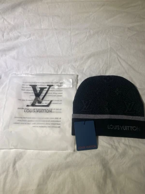 Svart grå mössa med logga - Snygg svart mössa med grå rand och broderad logga framtill. Mössan har diskret mönster och är stickad i mjukt material. Perfekt för dig som vill ha en exklusiv accessoar med stilren design.