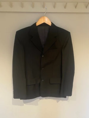 Svart kavaj med klassisk passform - Stilren svart kavaj med tre knappar framtill och klassisk krage. Två fickor med lock och en bröstficka ger en clean look. Materialet är polyester och kavajen har långärmade ärmar. Perfekt för dig som vill ha en enkel och snygg stil.