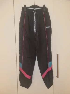 Svarta byxor från Adidas med färgdetaljer - Säljer ett par svarta byxor från Adidas med snygga rosa och turkosa detaljer längs benen