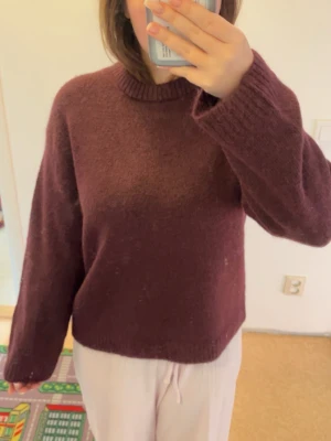 Vinröd stickad tröja - Säljer en mysig mörklila stickad tröja från Gina Tricot med rund hals och ribbade muddar. Tröjan har lång ärm och är tillverkad i en mjuk ullblandning som känns skön mot huden. Perfekt för kyliga dagar och enkel att matcha med jeans eller kjol.