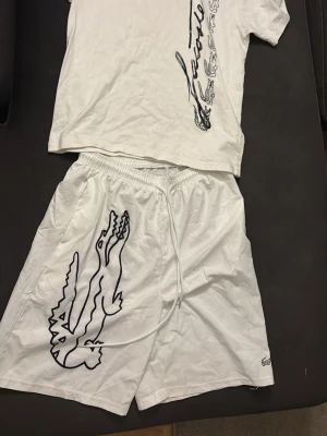 Vit  Lacoste dress med snörning - Säljer ett par vita shorts från Lacoste med stor svart krokodilprint på ena benet och klassisk snörning i midjan. Shortsens material känns lätt och luftigt, perfekt för varma dagar. Diskret Lacoste-logga broderad vid benslutet. Gul fläck vid bakkdel av shortsen