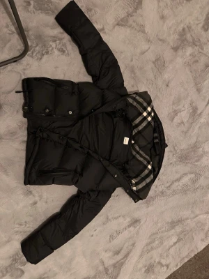 Svart Burberry pufferjacka med huva - Svart pufferjacka från Burberry med klassisk rutig huva i grått, vitt och svart. Jackan har dragkedja och knappar framtill, två sidofickor med dragkedja och en snygg Burberry-logga på ärmen. Perfekt för kalla dagar och riktigt stilren.  Den har ett jätte litet hål eller inte ens ett hål det är typ som att nån nål 📌 har gått in men man märker inget och den är köpt från modeza för 2800. Blir till en väst också!