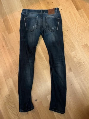 Adrian Hammond Nevada Slim jeans mörkblå - Mörkblå jeans från Adrian Hammond, modell Nevada Slim. Snygga slitningar och detaljer på benen, klassisk femficksdesign och knappgylf.  Perfekt för dig som gillar lite schystare och tightare jeans! Storlek 32 men små i storleken! Tveka inte att höra av dig vid frågor 