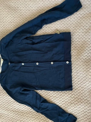 Marinblå barnkofta från POMPdeLUX - Snygg marinblå kofta från POMPdeLUX i storlek 122/128. Koftan har långa ärmar, ribbade muddar och vita knappar framtill. Materialet är mjukt och stickat, perfekt för lager-på-lager. Klassisk och enkel design som funkar till många olika stilar.