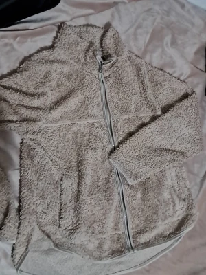 Beige teddykofta från 157, storlek S - Mysig beige teddykofta från 157 med hel dragkedja framtill och två sidofickor. Jackan har hög krage och är tillverkad i fluffigt fleece-material som ger en skön och avslappnad vibe. Perfekt för kyliga dagar när du vill vara både bekväm och snygg.