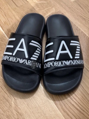  Emporio Armani sandaler Storlek 40 - Svarta sandaler från Emporio Armani med stor vit logga 'EA7' och text på ovansidan. Snygg och enkel design med platt sula och öppen tå. Tillverkade i syntetmaterial med bekväm fotbädd. Perfekt för chill dagar och enkel att slänga på sig.