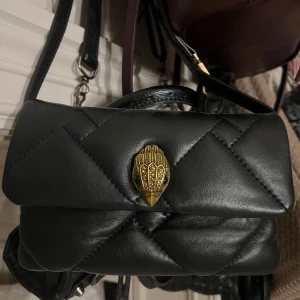 Svart quiltad axelväska Kurt Geiger - Snygg svart quiltad axelväska från Kurt Geiger med guldfärgad fågeldekor på locket och justerbar axelrem. Väskan är tillverkad i mjukt skinn och har en elegant metallbricka med logga på baksidan. Perfekt för dig som gillar stilrena detaljer och lyxig känsla.