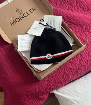 Svart Moncler mössa med logga - Snygg svart mössa från Moncler med klassisk logga framtill och rödvit rand längst ner. Perfekt accessoar för kyliga dagar och ger en sportig touch till din outfit.