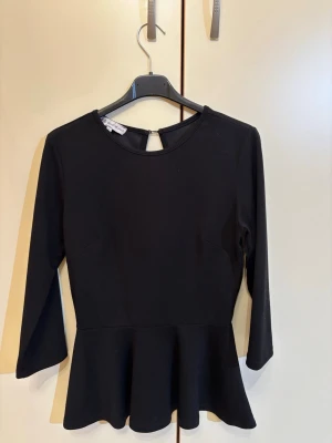 Svart peplumtop från Zic Zac - Säljer en svart peplumtop från Zic Zac med rund halsringning och snygg öppning bak. Toppen har lång ärm och är tillverkad i ett mjukt syntetmaterial. Den har en figurnära passform upptill och en volang nedtill som ger en cool siluett.