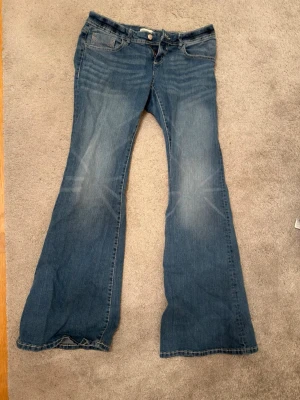 Blå bootcut jeans med kontrastsömmar - Snygga blå bootcut jeans från Gina med klassisk femficksdesign och kontrastsömmar på bakfickorna. Jeansen har låg midja och är tillverkade i mjukt denim-material. Passar dig som gillar retrovibbar och breda ben. Jeansen är aldrig använda och köpta för 400kr, säljer för 250