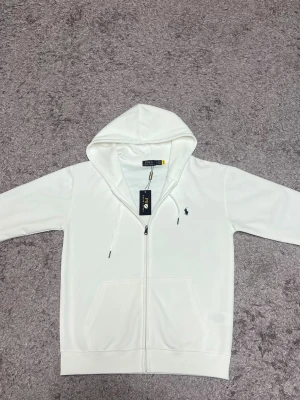 Polo Ralph lauren tröja med dragkedja - Hej!  Jag säljer en vit Ralph lauren kofta. Den är helt ny och har bara testat den en gång, den har väldigt bra kvalité och passar för alla årstider. Jag säljer den eftersom jag fick den i present och den var lite för stor. Den passar både M och L men hade nog sutit bättre på L.        Pris kan diskuteras!