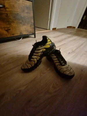 Nike Air Max Plus TN gul/svart - Nike Air Max Plus TN sneakers i en fet gul och svart färgkombination med vågiga svarta detaljer på ovandelen. Skorna har en robust svart sula och meshmaterial som andas. Perfekta för dig som vill sticka ut med en unik och sportig look.