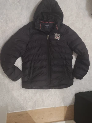 Svart pufferjacka från Vinson Polo Club - Svart pufferjacka med huva från Vinson Polo Club. Jackan har broderad logga på bröstet, två fickor med dragkedja och snygga röda detaljer vid kragen. Perfekt för kalla dagar och har en quiltad look som ger extra stilpoäng. Det står L men den sitter som M, ena fixan är lite trasig och dragkedjan nere från början är lite trasig men funkar at använda såklart!