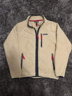 Patagonia fleece  - Säljer en beige fleecejacka från Patagonia i storlek S. Jackan har marinblå detaljer, röda dragkedjor och hög krage. Tre fickor med dragkedja framtill och mjukt, mysigt material som håller dig varm under kyliga dagar.