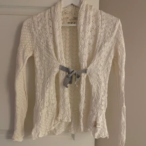 Vit kofta med rosett från Odd Molly - Säljer en vit, långärmad kofta från Odd Molly med vacker spetsstickning och grå rosett framtill. Koftan har ett öppet omlott-utförande och fina detaljer i mönstret. Supersöt kofta!!