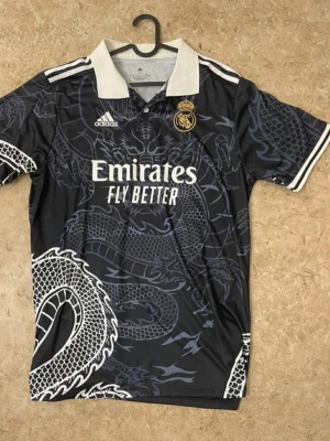 Real Madrid x Adidas svart draktröja - Säljer en svart Real Madrid x Adidas matchtröja med stort drakmönster i grått och vitt över hela plagget. Tröjan har vit krage, vita ränder på axlarna och Real Madrid-logga på bröstet. Materialet är lätt polyester och trycket är riktigt detaljerat.