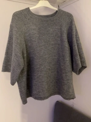 Grå stickad tröja från 157, S - Mysig grå stickad tröja från 157 i storlek S. Tröjan har rund hals, korta ärmar och en loose passform som ger en chill vibe. Perfekt att slänga på sig när det är lite kyligt ute. Enkel och stilren design som funkar till jeans eller kjol.