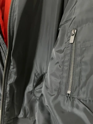 Svart bomberjacka Black Label - Snygg svart bomberjacka från The Black Label Collection med orange foder. Jackan har klassisk ribbad krage, dragkedja framtill och fickor med dragkedja på ärmen. Materialet är glansig polyester som ger en cool vibe. Perfekt för dig som gillar streetstyle.