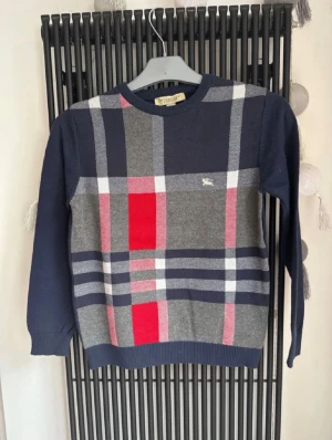 Rutig tröja från Burberry London - Snygg långärmad tröja från Burberry London med klassiskt rutigt mönster i marinblått, grått, rött och vitt. Tröjan har rund hals och ribbade muddar. Materialet är 100% bomull, vilket gör den mjuk och skön att bära. Perfekt för dig som gillar stilrena designer med ikonisk touch.