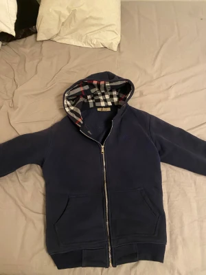 Burberry kofta äkta! - Säljer en marinblå hoodie från Burberry i storlek S. Tröjan har dragkedja, huva med klassiskt rutigt Burberry-mönster i blått, vitt, svart och rött, samt två fickor framtill. Materialet är mjukt och fleece-liknande på insidan, perfekt för kyliga dagar. Koftan är äkta och det finns inte skador på den för att jag har inte använt den så mycket alls 