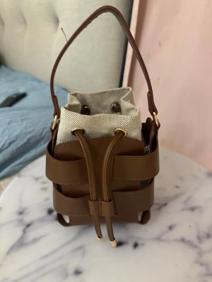 Brun handväska med snörning - Snygg handväska i brun skinnimitation med coolt flätad design och detaljer i guld. Väskan har en beige tyginsats med snörning upptill och ett stadigt handtag. Perfekt storlek för det viktigaste och ger en trendig touch till din outfit.