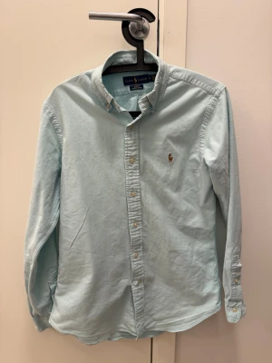 Ljusblå skjorta från Ralph Lauren M - Snygg ljusblå skjorta från Ralph Lauren i slim fit med klassisk button down-krage och broderad logga på bröstet. Skjortan är tillverkad i stretchig bomull och har vita knappar framtill. Perfekt för dig som gillar stilrena och tidlösa plagg.