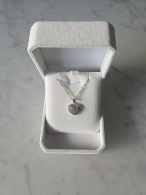 Halsband med glittrigt hjärthänge - Snyggt halsband i silverfärg med ett hjärtformat hänge som är täckt av glittrande stenar. Kedjan är tunn och stilren, och hänget har ett extra litet hjärta i mitten för en gullig touch. Perfekt för dig som gillar romantiska detaljer och bling.