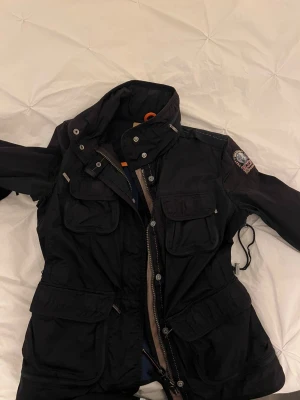 Svart Parajumpers parkajacka - Svart parkajacka från Parajumpers. Jackan har dragkedja och knappar framtill, samt justerbar midja med snörning. Jackan är äkta 
